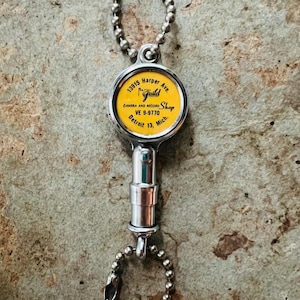 May include: A silver-toned metal retractable badge reel featuring a circular yellow insert with black text that reads 13915 Harper Ave. Guild Camera and Record Shop VE 9-9770 Detroit 13, Mich. The device includes two silver ball-chain attachments.
