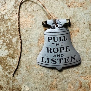 May include: A bell-shaped sign with the text "PULL THE ROPE AND LISTEN" in black. The sign is light blue with a brown rope attached to the top. The background is a textured, mottled brown.