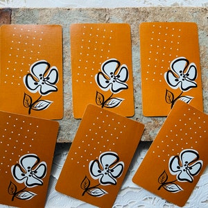 May include: Six rectangular playing cards featuring a mustard yellow background with a white and black graphic flower illustration and a pattern of small white dots in the upper left corner. Each card displays the same floral design.