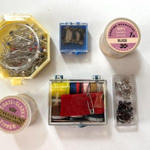 May include: A collection of sewing supplies including a yellow hexagonal container of multi-colored pins, a small blue box of metal hooks, a 7-inch black zipper spool, a clear box of assorted thread spools, a Coats & Clark's zipper, and a small clear case of fasteners.