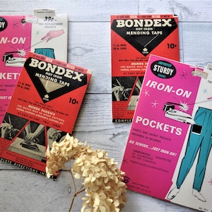 May include: A collection of vintage sewing supplies, including pink and white "Iron-On Pockets" and red and black "Bondex Hot Iron Mending Tape" packages. The packages feature illustrations of clothing and instructions. The items are displayed on a light-colored surface.