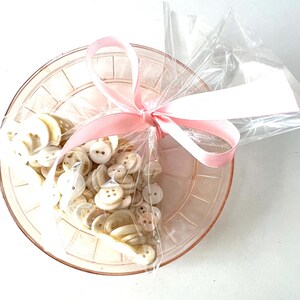 May include: A collection of off-white buttons in a clear cellophane bag, tied with a light pink ribbon. The bag sits inside a pale pink, decorative dish. The buttons vary in size and style, with two or four holes.