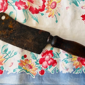 May include: An antique meat cleaver with a dark, aged blade and a wooden handle. The cleaver has a hole near the top and shows signs of wear. It rests on a floral patterned tablecloth with red, yellow, and blue flowers.