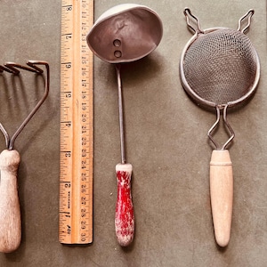 May include: A collection of vintage kitchen utensils, including a potato masher, a ladle, and a strainer. The masher has a wooden handle and metal tines. The ladle has a red and white speckled handle. The strainer has a wooden handle and a wire mesh.