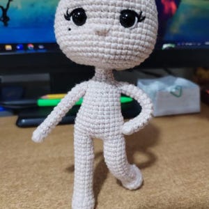 May include: A crocheted doll with a round head, black eyes, and a small nose. The doll has a light beige color and is posed with one arm raised and one leg forward. The doll is standing on a brown surface.