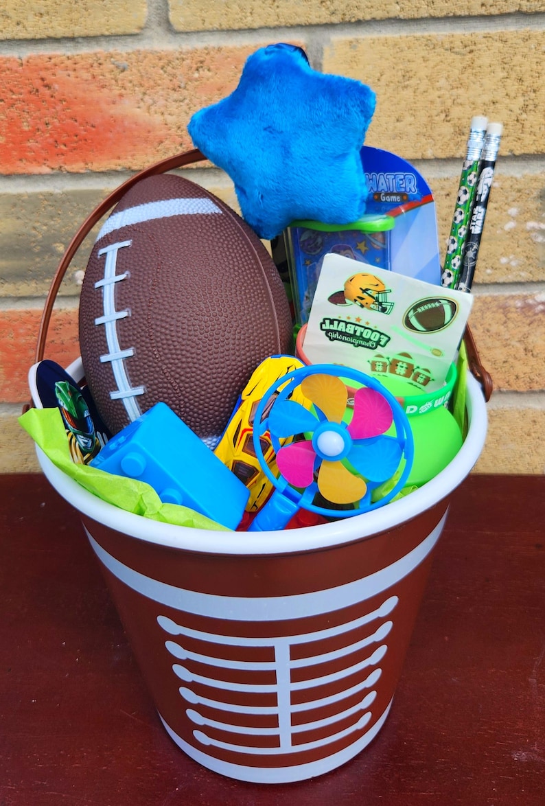 May include: A brown and white football-themed plastic bucket filled with a brown football, a blue plush star, two pencils with football patterns, a handheld blue and multicoloured pinwheel, a yellow toy car, and a football sticker sheet.