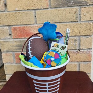 May include: A brown plastic bucket with white laces printed on the front, filled with a brown football, a blue plush star, two pencils with football patterns, a small blue pinwheel, a blue building block, and a sheet of football stickers.