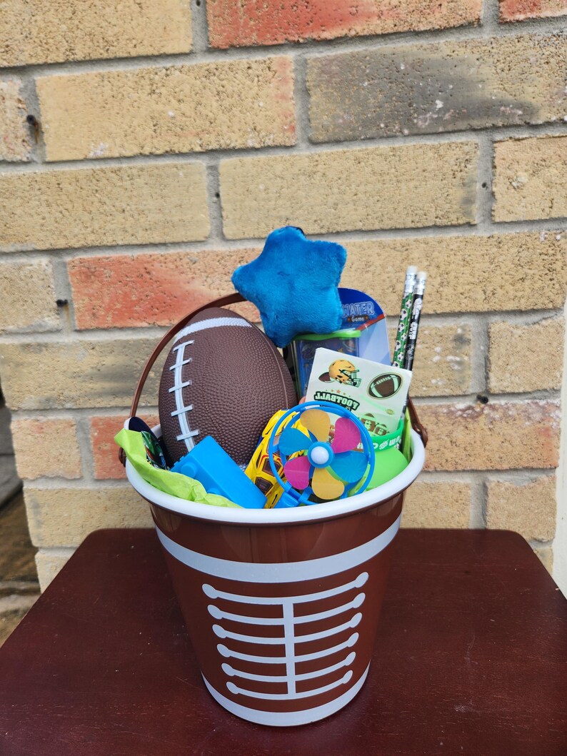 May include: A brown plastic bucket with white laces printed on the front, filled with a brown football, a blue plush star, two pencils with football patterns, a small blue pinwheel, a blue building block, and a sheet of football stickers.