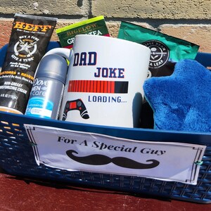 May include: A blue basket filled with gifts, including a mug that says "DAD JOKE LOADING...", a blue star-shaped plush, and toiletries. The basket has a sign that reads "For A Special Guy" with a mustache graphic.