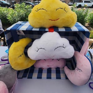 Könnte beinhalten: Vier Plüschtiere in Form von Wolken und einem Halbmond. Die Wolken sind weiß, gelb und rosa mit gestickten Augen und Lächeln. Der Mond ist gelb mit gestickten Augen und einem Lächeln. Die Spielzeuge sind in einer blau-weiß karierten Schachtel angeordnet.