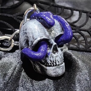 Pode incluir: Um pingente de caveira prateada com uma cobra roxa enrolada. A caveira tem detalhes e é presa a uma corrente prateada. O pingente é exibido em um tecido preto.