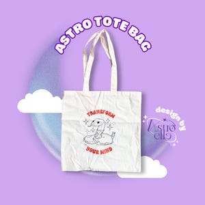 May include: White canvas tote bag with the text "ASTRO TOTE BAG" in a purple arc at the top. The bag features a graphic of a snake with the words "TRANSFORM YOUR MIND" in red. The design is by "Astro Ella".