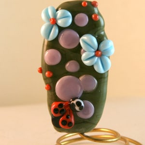 Op de afbeelding: Een groene glazen kraal met een stippenpatroon, versierd met twee blauwe bloemen met rode centra en een rood lieveheersbeestje met zwarte stippen.