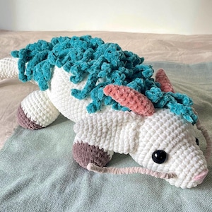 May include: A white crocheted dragon with teal and pink accents. The dragon has a long tail and a large head with black eyes. The dragon is lying on a light blue blanket.