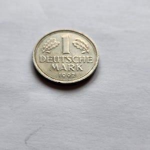 Puede incluir: Una moneda de plata de 1 Deutsche Mark de 1992. La moneda presenta el texto "DEUTSCHE MARK" y el número "1" en el centro, con hojas de roble a ambos lados. La moneda tiene un borde estriado.