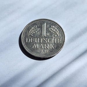 May include: A silver Deutsche Mark coin from 1991. The coin features the number 1 in the center, with the words "DEUTSCHE MARK" below it. The coin is surrounded by a decorative border and two sprigs.