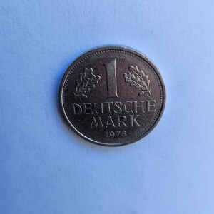 Puede incluir: Una moneda plateada con el número 1, la inscripción "DEUTSCHE MARK" y el año 1978. La moneda tiene un borde detallado y está sobre un fondo azul claro. La moneda presenta una ramita de hojas a cada lado del número 1.