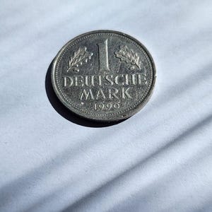 May include: A silver Deutsche Mark coin from 1990. The coin features the number 1, the words "DEUTSCHE MARK", and the year 1990. The coin has a detailed design with leaves on either side of the number 1.