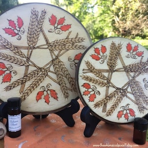 May include: Two wooden decorative pieces with a pentagram design, featuring wheat and red leaves. The design is burned into the wood. Small bottles of essential oil are in front of the pieces. The text on the bottles reads "Autumn Blend".