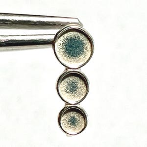 May include: A silver pendant with three circular settings, each containing a textured, light blue-green material. The pendant is held by a pair of tweezers against a white background. The design is simple and elegant.