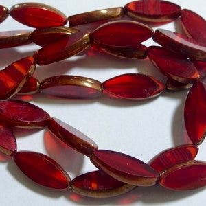 May include: A string of oval beads in a deep red colour with a metallic gold-coloured rim. The beads are strung together with a thin, clear string, creating a necklace or bracelet component. The beads have a glossy finish.