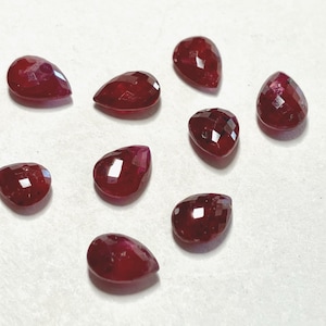 May include: Nine faceted, teardrop-shaped, deep red gemstones are scattered on a white surface. The gems have a polished, reflective surface, catching the light and displaying a range of crimson hues. These gemstones are suitable for jewelry making.