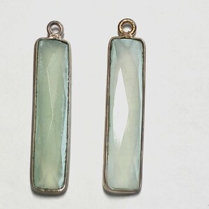 May include: Two rectangular pendants featuring faceted pale green stones set in metallic frames. Each pendant has a small circular loop at the top for attachment. The stones have a translucent quality with visible geometric facets on the surface.