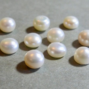 May include: A collection of ten lustrous, round pearls. Each pearl has a creamy white color with a subtle iridescent sheen. The pearls are scattered on a neutral gray surface, showcasing their smooth, spherical shape.