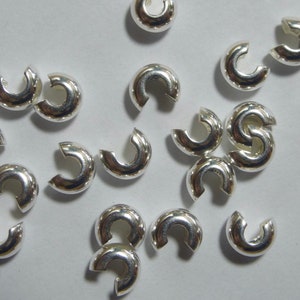 May include: A collection of shiny, silver-colored C-shaped jewelry clasps. These small, curved components are designed to connect jewelry pieces. The clasps are scattered on a light-colored surface, reflecting light.