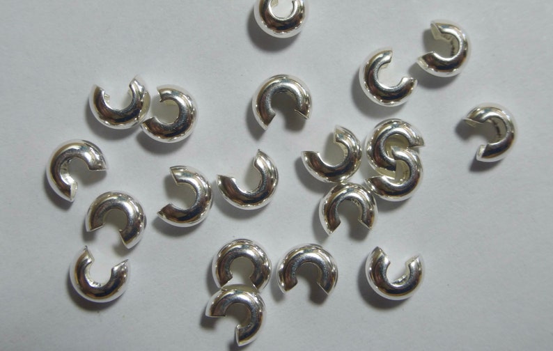 May include: A collection of shiny, silver-colored C-shaped jewelry clasps. These small, curved components are designed to connect jewelry pieces. The clasps are scattered on a light-colored surface, reflecting light.