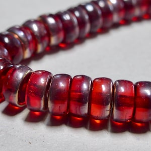 May include: Close-up of a strand of red glass beads. The beads are cylindrical with a slightly rounded shape and a metallic gold-colored band around the middle. The beads are strung together on a thin, clear string.