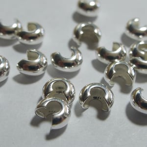 May include: A collection of shiny, silver-colored crimp beads. These small, C-shaped beads are designed for jewelry making and crafting. The beads are scattered across a light-colored surface, reflecting light.