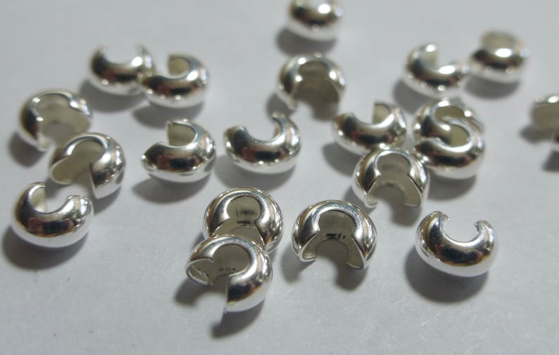 May include: A collection of shiny, silver-colored crimp beads. These small, C-shaped beads are designed for jewelry making and crafting. The beads are scattered across a light-colored surface, reflecting light.