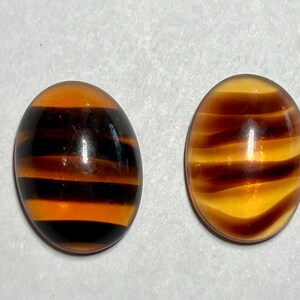 May include: Two oval-shaped cabochons with a striped pattern. One features alternating bands of black and orange, while the other has bands of brown and yellow. These polished stones are likely used for jewelry making.
