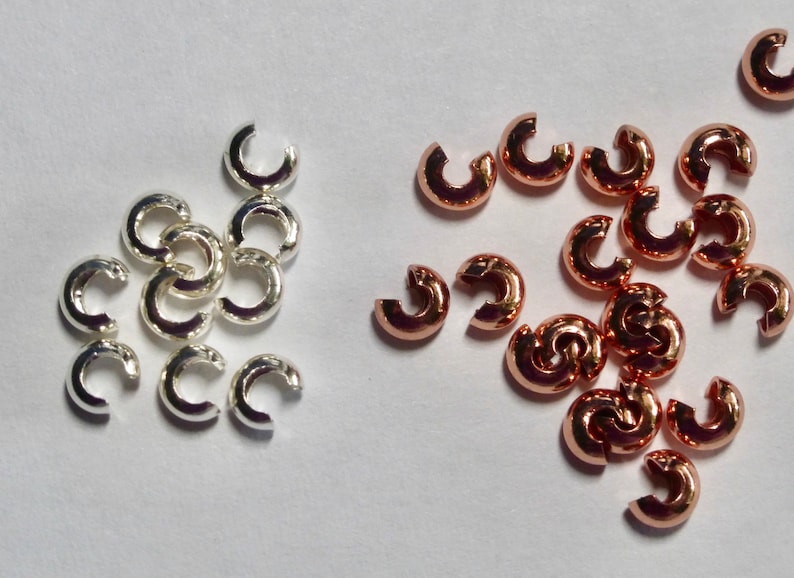 May include: A collection of silver and copper-colored C-shaped crimp beads. The beads are small, with a smooth, rounded appearance. They are used in jewelry making to secure clasps and other components.