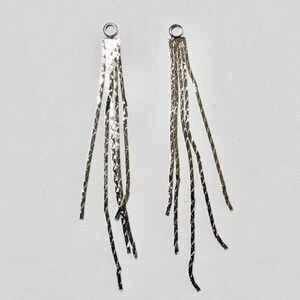 May include: Two silver-colored, dangling earrings. Each earring features a rectangular top with a small loop and multiple strands of thin, textured metal hanging down. The earrings are set against a white background.