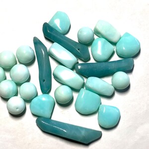 May include: A collection of loose, polished gemstones in various shades of light blue, turquoise, and teal. The assortment includes smooth spherical beads, irregular flat shapes, and elongated, tapered pieces with a glossy, reflective surface finish.
