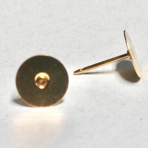 May include: Two gold-colored earring posts with flat, round backs. One post is upright, showing the flat disc and the post. The other post is lying down, showing the back of the disc and the post. The earrings are on a white surface.