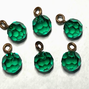 May include: Six faceted emerald green glass beads with small bronze-colored loops for stringing. The beads have a round shape with multiple flat surfaces, reflecting light. These beads are suitable for jewelry making.