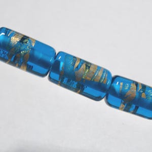 May include: Three blue glass beads with gold leaf accents are strung on a wire. The beads are cylindrical with rounded edges. The gold leaf creates a swirling pattern within the transparent blue glass.