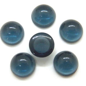 May include: Six blue-green glass cabochons, arranged on a white surface. Five are rounded, domed shapes, and one is a flat-backed cabochon. The glass has a glossy finish, reflecting light. These are suitable for jewelry making or craft projects.