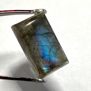 May include: A rectangular labradorite gemstone held by tweezers. The stone has a polished surface with iridescent blue and green flashes. The gem is a mix of gray, brown, and blue hues, with a rectangular shape.