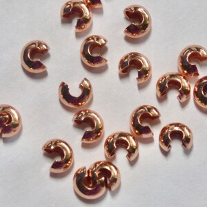 May include: A collection of rose gold-colored crimp beads, scattered on a white surface. These small, C-shaped beads are designed for jewelry making, with a smooth, metallic finish. They are used to secure clasps and other components.