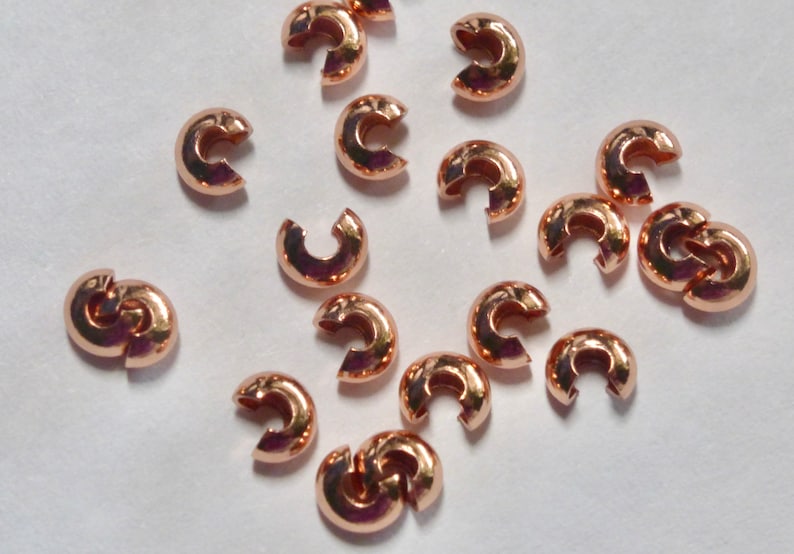 May include: A collection of rose gold-colored crimp beads, scattered on a white surface. These small, C-shaped beads are designed for jewelry making, with a smooth, metallic finish. They are used to secure clasps and other components.