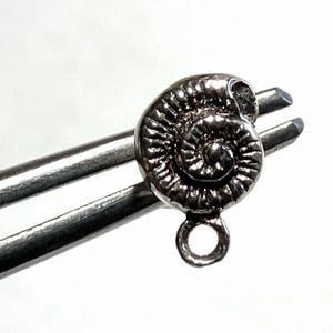 May include: A small, silver-toned metal charm shaped like a coiled ammonite fossil. The charm features a detailed, textured spiral pattern on its surface and includes a circular jump ring at the bottom for attachment to jewelry or other items.