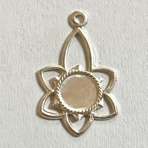 May include: A silver-colored pendant with a floral design. The pendant features a central circular setting surrounded by a rope-like border and six petal-shaped elements. A loop is at the top for attaching to a chain or cord.