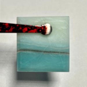 May include: A square-shaped piece of polished blue and white stone with a distinct horizontal grey vein running across the center. The stone is being held by a pair of black tweezers featuring a red and white speckled pattern on the handle.