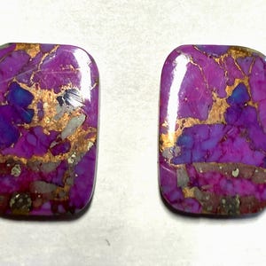 May include: Two rectangular cabochons with rounded corners, featuring a vibrant purple base color with intricate copper-colored veining and scattered grey and dark inclusions throughout the polished, smooth surface of the stones.