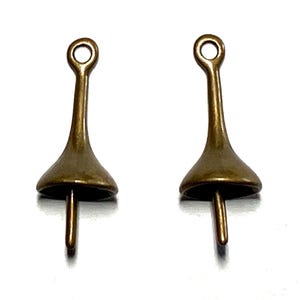 May include: Two antique brass-colored metal charms. Each charm has a flared, bell-shaped body with a small loop at the top and a pin extending from the bottom. The charms are displayed against a white background.