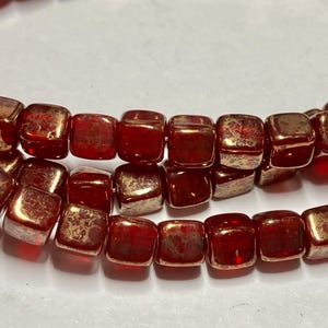 May include: Close-up of two strands of square, red beads with a metallic gold finish on the edges. The beads are strung together, creating a textured and reflective surface. The beads are likely for jewelry making.
